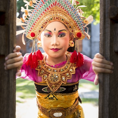 En 6 år gammel søt, asiatisk liten jente har på seg en tradisjonell Bali-drakt og stirrer gjennom en treskåret dør på et tempelsted. Barn lærer i veldig ung alder den eldgamle kunsten Ramayana-dans og andre hindu-danser, som vanligvis foregår i tempelskoler.