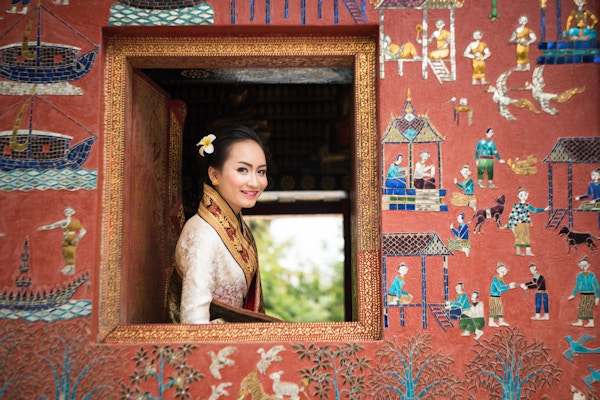 Foto av en vakker Laos-dame i tradisjonelt kostyme som smiler og ser ut av et vindu fra et tempel (Wat Xieng Thong) i Luang Prabang. Fotografiets fokus ligger på ansiktet hennes med mykt fotofokus på området rundt.
