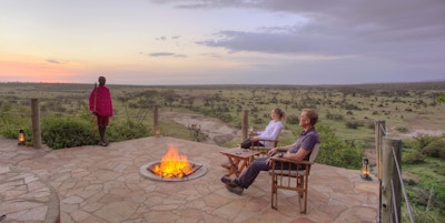 Par sitter på utsiktsplattform med bålpanne og ser utover savannen, masai kledd i rødt og rosa står ved siden av.