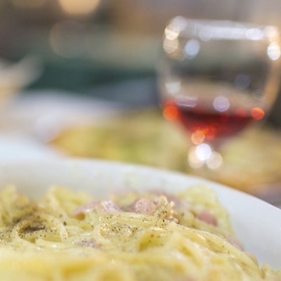 Nærbilde av en bolle med spaghetti a la carbonara. Et glass rødvin og brød er synlige i bakgrunnen.