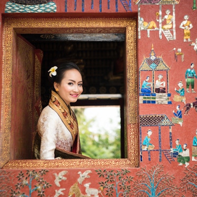 Foto av en vakker Laos-dame i tradisjonelt kostyme som smiler, og ser ut av et vindu fra et tempel (Wat Xieng Thong) i Luang Prabang. Fokus på ansiktet hennes og mykt fokus på området rundt.