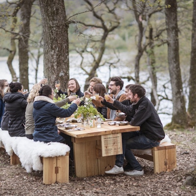 Gruppe menneske sitter ved utendørs langbord om vpren og spiser og drikker.