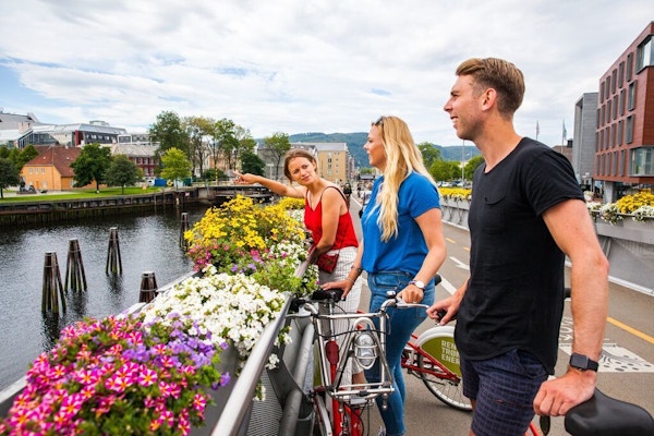 Tre mennesker med sykler står langs elva med sommerblomster.