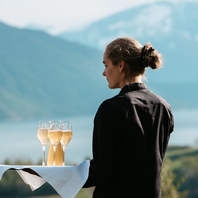 Servitør som holder ett brett med vin i fokus. Fjord og sjø i bakgrunnen