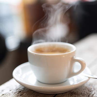 Damp stiger opp fra en hvit kopp varm kaffe med en skje på en tallerken over et trebord i kafeen. Nærbilde av en forfriskende varm kopp kaffe på en kafé.