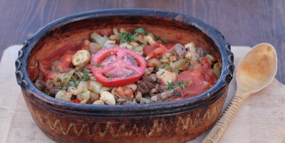 Terrakottaskål fylt med kjøttgryte med sopp, løk og urter toppet med en halv tomat servert på en trefjøl med en treskje på siden