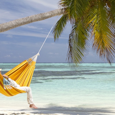 En ung mann iført stråhatt slapper av i en oransje hengekøye som er hengt på et kokosnøttpalm. Den hvite sandstranden og fargen på vannet er turkis og er en idyllisk og unik beliggenhet på Maldivene.