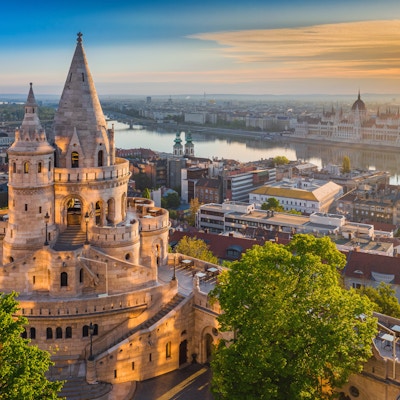 Budapest, Ungarn - Vakker gylden sommer soloppgang med tårnet til Fisherman's Bastion og grønne trær. Ungarns parlament og Donau i bakgrunnen. Blå himmel.