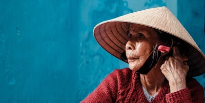 Kvinne i rød cardigan og tradisjonell vietnamesisk hatt sitter ved en blå vegg