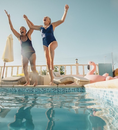 To eldre kvinner som holder hender og hopper ut i et svømmebasseng. De er glade og har det gøy.