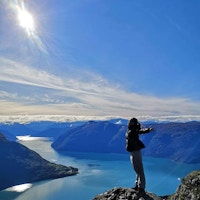 Kvinne med armene utstrakt mot sola på toppen av fjellet med fjord og fjell i bakgrunnen.