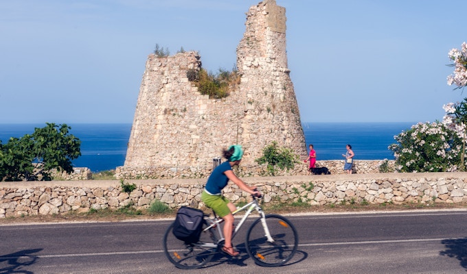 En dame sykler på veien langs kysten foran et gammelt murtårn i Puglia