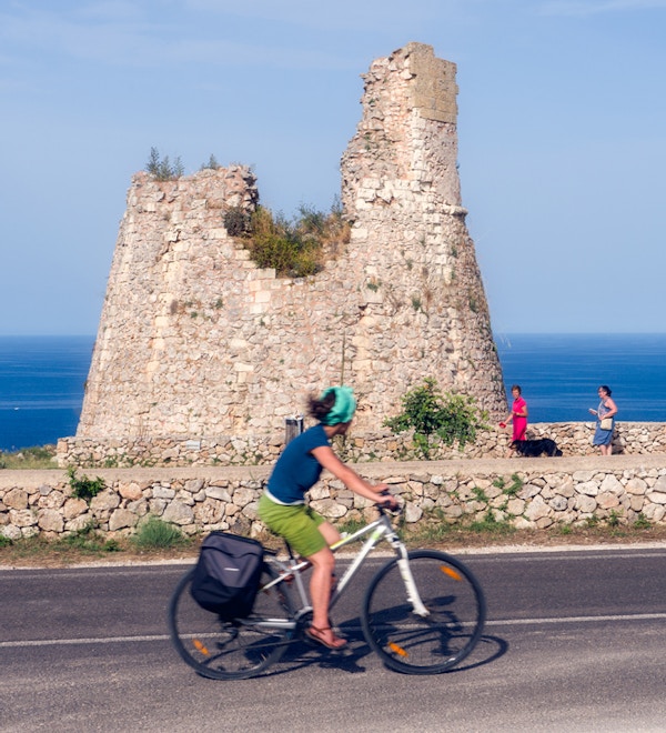 En dame sykler på veien langs kysten foran et gammelt murtårn i Puglia
