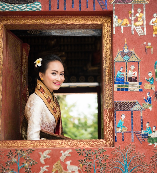 Foto av en vakker Laos-dame i tradisjonelt kostyme som smiler, og ser ut av et vindu fra et tempel (Wat Xieng Thong) i Luang Prabang. Fokus på ansiktet hennes og mykt fokus på området rundt.