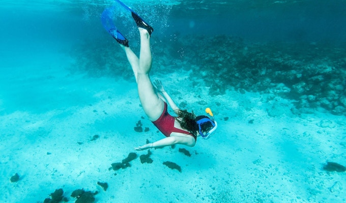 Kvinne dykker ned mot sandbunnen med snorkel, iført rød badedrakt og blått utstyr