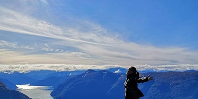 Kvinne med armene utstrakt mot sola på toppen av fjellet med fjord og fjell i bakgrunnen.