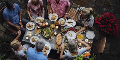 En gruppe voksne venner sitter rundt et utendørs spisebord og spiser og drikker. De snakker alle lykkelig og gleder seg over hverandres selskap. Bildet er tatt ovenfra, i Toscana, Italia.