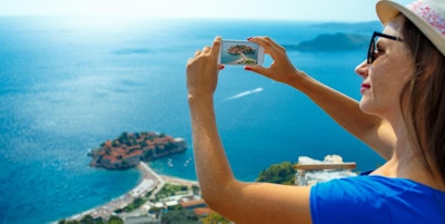 En kvinne med solhatt som tar bilder ved byen Sveti Stefan i Montenegro, Balkan