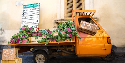 En søt oransje vintagebil som selger lilla blomkål på en gate i Sicilia, Italia. Gul vegg, skodder og veiskilt i bakgrunnen.