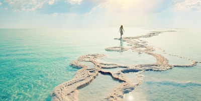 Silhuett av en ung kvinne som går på salt havkyst ved Dødehavet ved soloppgang mot solen