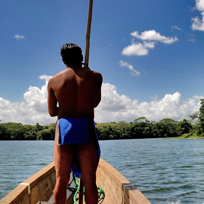 Sentralamerikanske guide som fører båten. Elv, blå himmel, skyer og grønn skog.