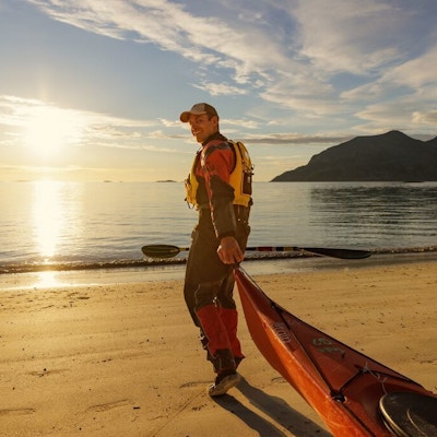 Mann på strand drar kajakk ut på havet med gyllen sol.