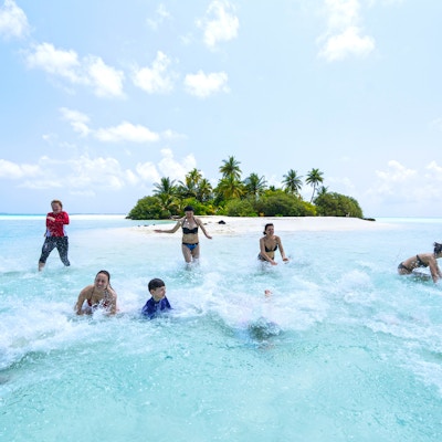 Frontutsikt til vakre unge, middelaldrende og eldre mennesker som har det gøy mens de løper og faller i grunt vann på sandstranden på den lille øya med palmer i bakgrunnen. De har komiske ansiktsuttrykk og er omgitt av turkis vann, horisonten og en fin blå himmel med få skyer. Det er ikke langt fra Maafushi-øya, Maldivene, og er en tropisk paradisøy, et av stedene du kan besøke fra Maafushi-øya på dagstur.